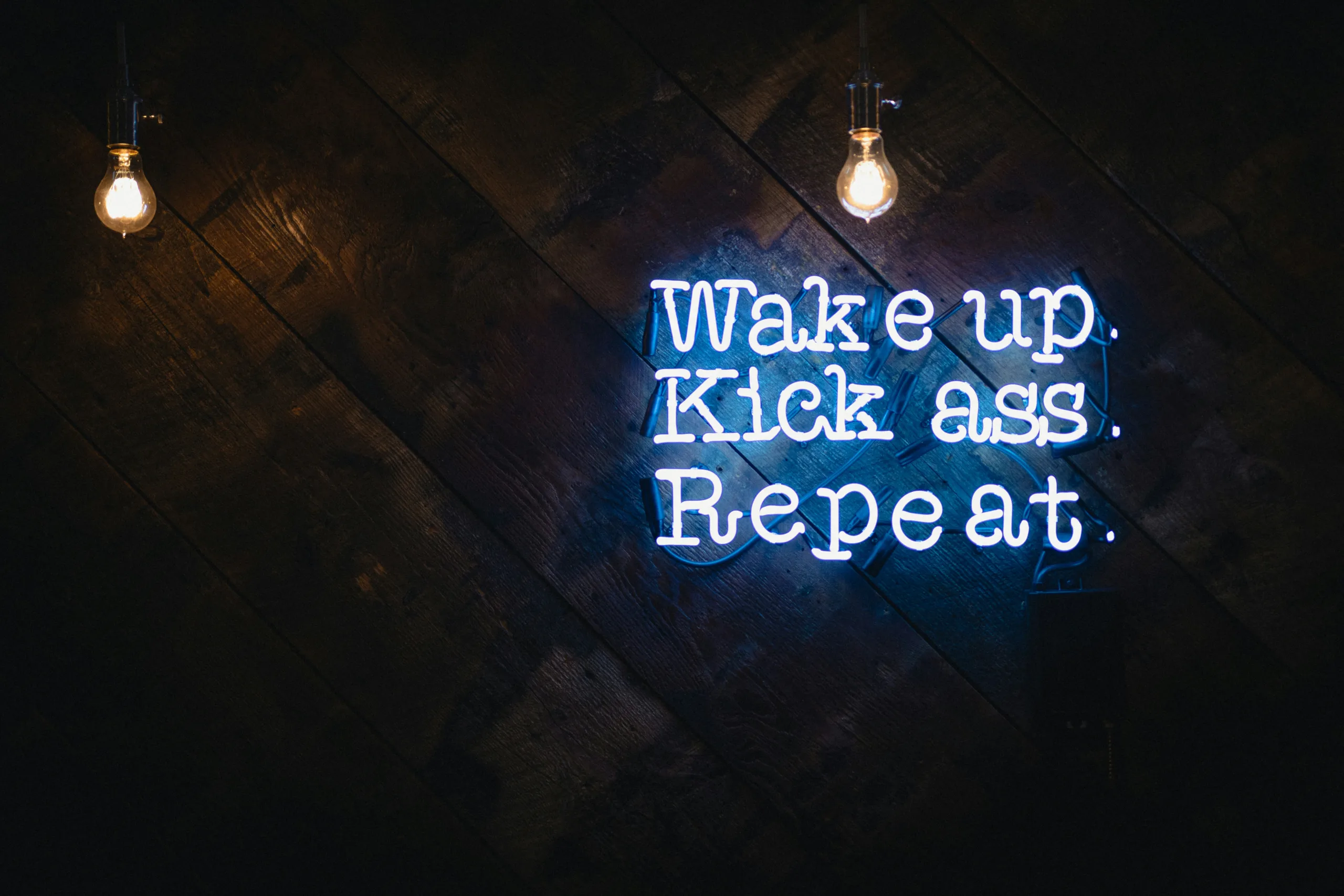 Gym motivation Neon sign reading "Wake up. Kick ass. Repeat." with two light bulbs glowing against a dark wooden background.